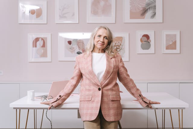 Elderly woman standing in front of desk and wall of pictures.